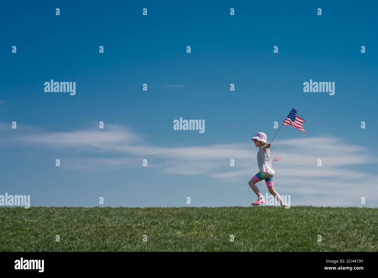 young girl runs across a grass hill waving 2 American flags Stock Photo