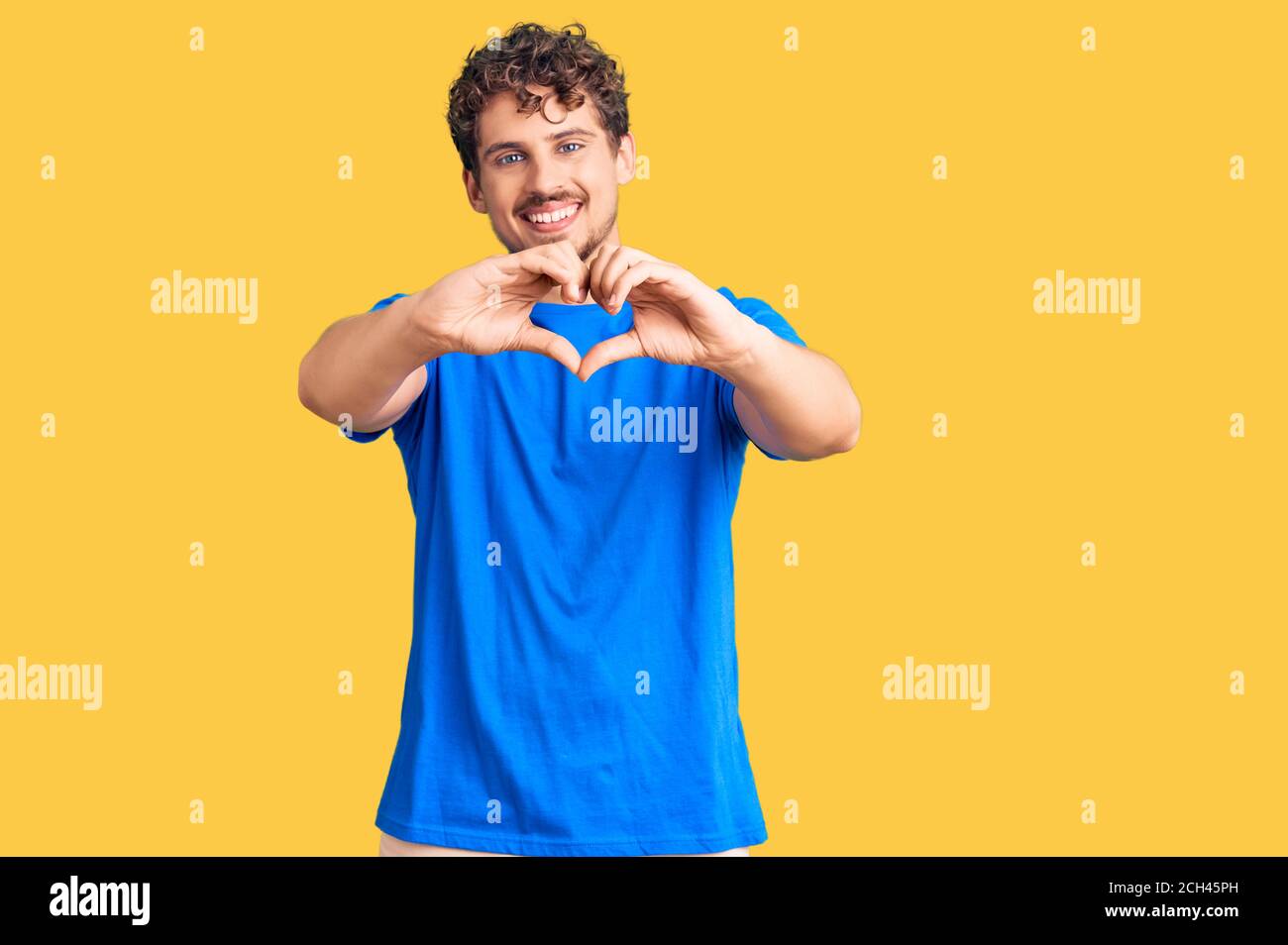 Young handsome man with curly hair wearing casual clothes smiling in love doing heart symbol shape with hands. romantic concept. Stock Photo