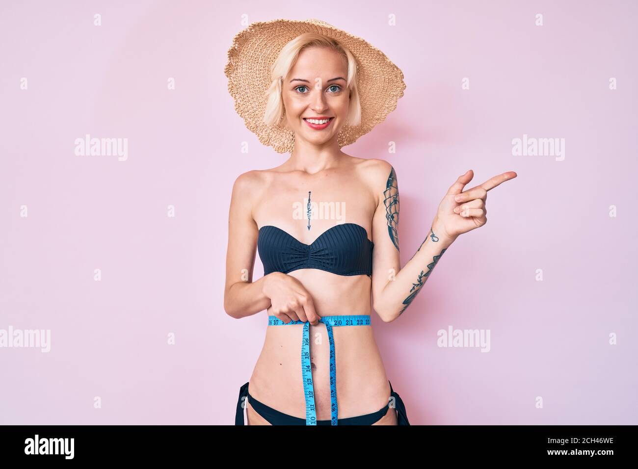 Young blonde woman with tattoo wearing bikini using tape measure smiling  happy pointing with hand and finger to the side Stock Photo - Alamy
