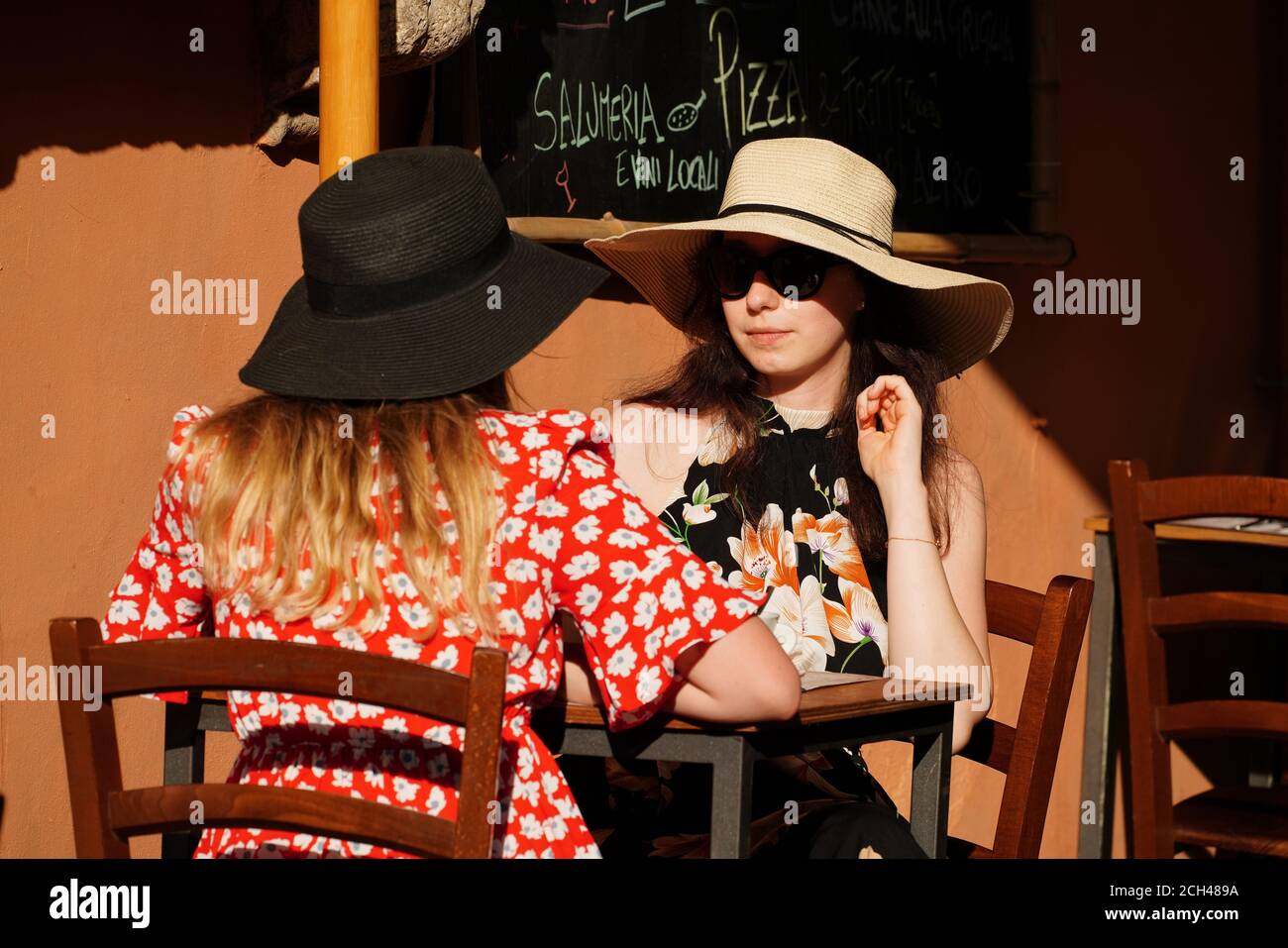Two Attractive, Stylish Young Women Wearing Wide-Brimmed Hats and Sunglasses Have Lunch Outside a Typical Restaurant in Famous Tourist Spot Trastevere Stock Photo