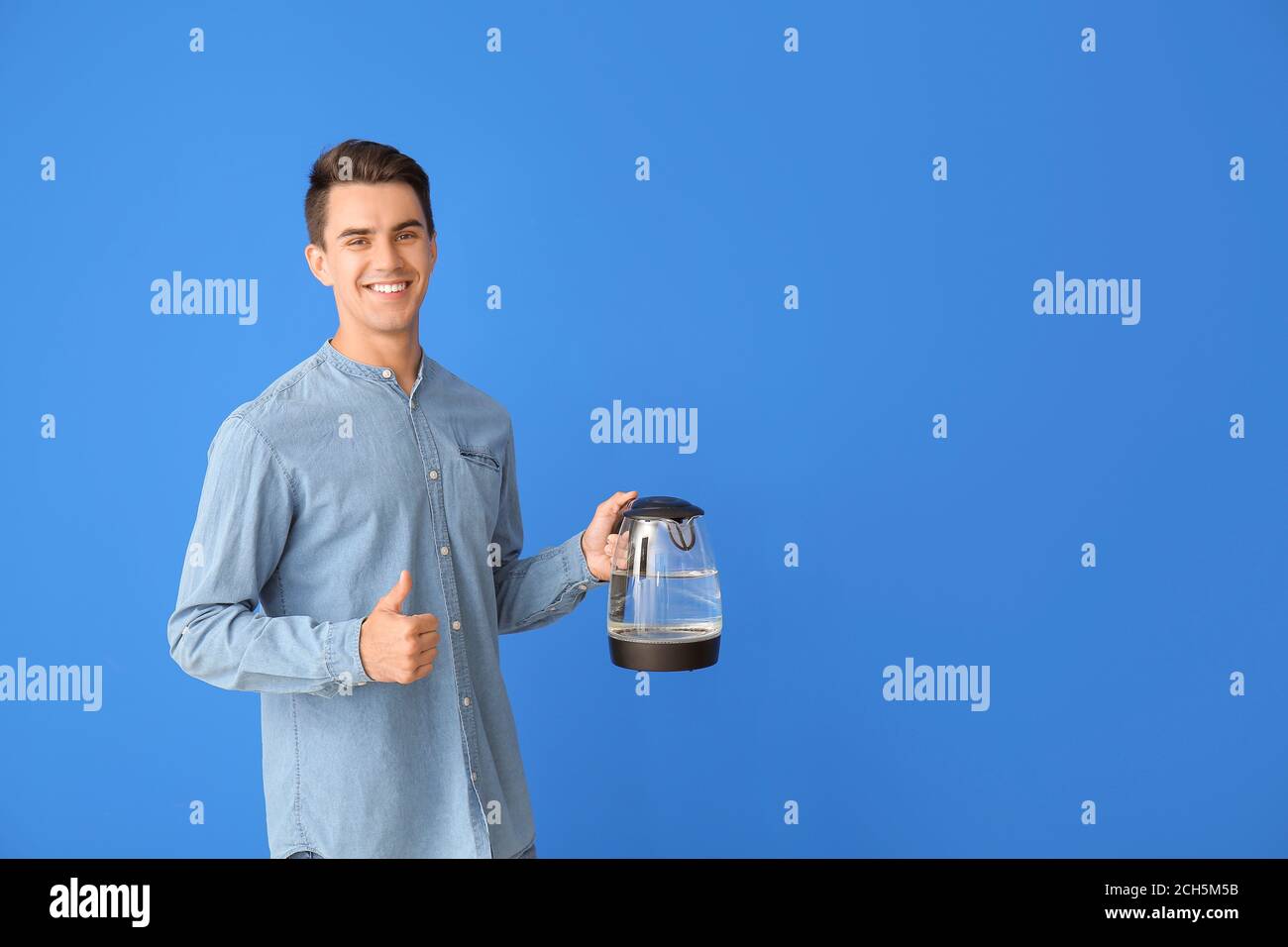 Young man with electric kettle showing thumb-up on color background ...