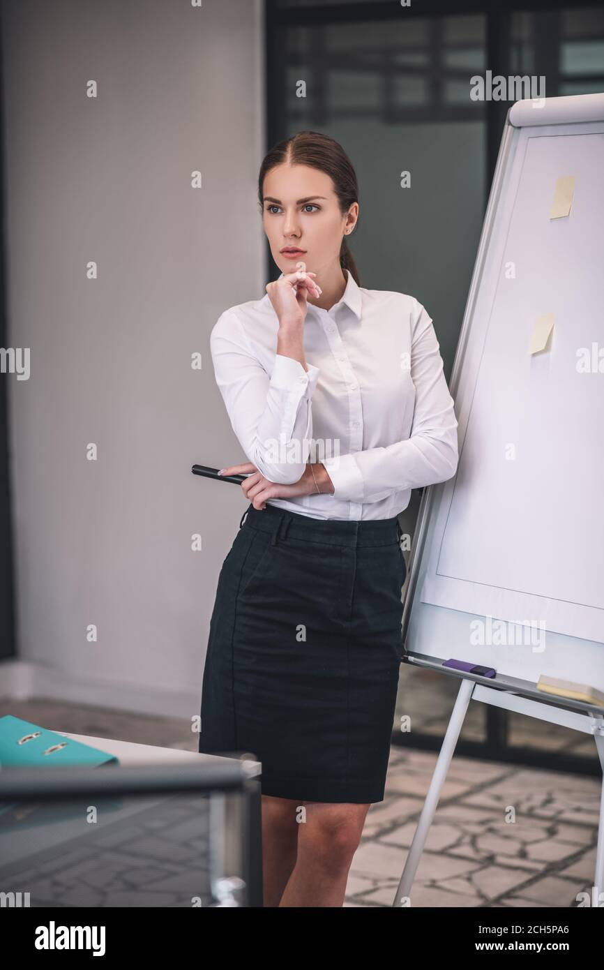 Brown-haired female in white shirt leaning on flipchart, thinking Stock Photo