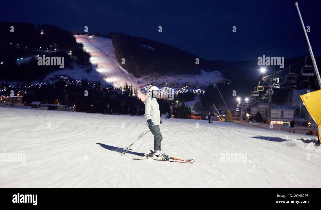 Side View Of Female Unrecognizable Skier In Ski Mask After Skiing Day Standing On Skis Evening Pastime At Resort Fairy Illuminated Mountain Countryside View And Chairlift In Darkness On Background Stock Photo
