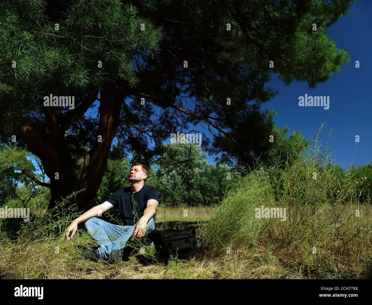 Male tourist meditates while sitting under a tree in the forest. Stock Photo