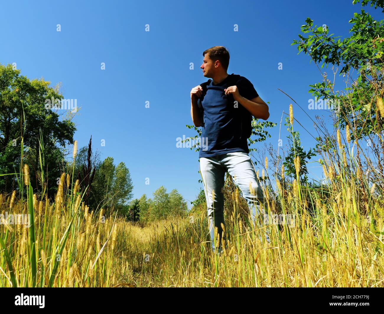 A man tourist with a backpack looks around while standing on a path in summer nature. Stock Photo