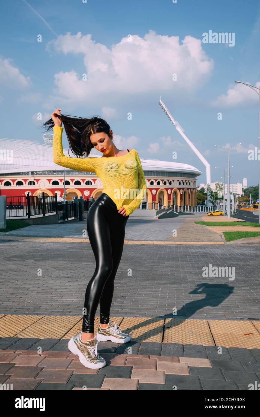 Pretty girl with long dark hair is wearing a bright yellow sweater, shiny, tight dark leggings & golden sneakers; in the background is a stadium Stock Photo