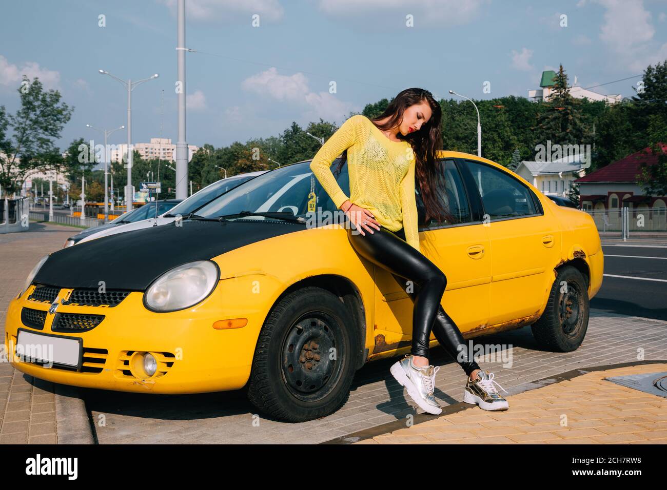 Pretty girl with long dark hair is wearing a bright yellow sweater, shiny, tight dark leggings and golden sneakers; she leans against a yellow car Stock Photo