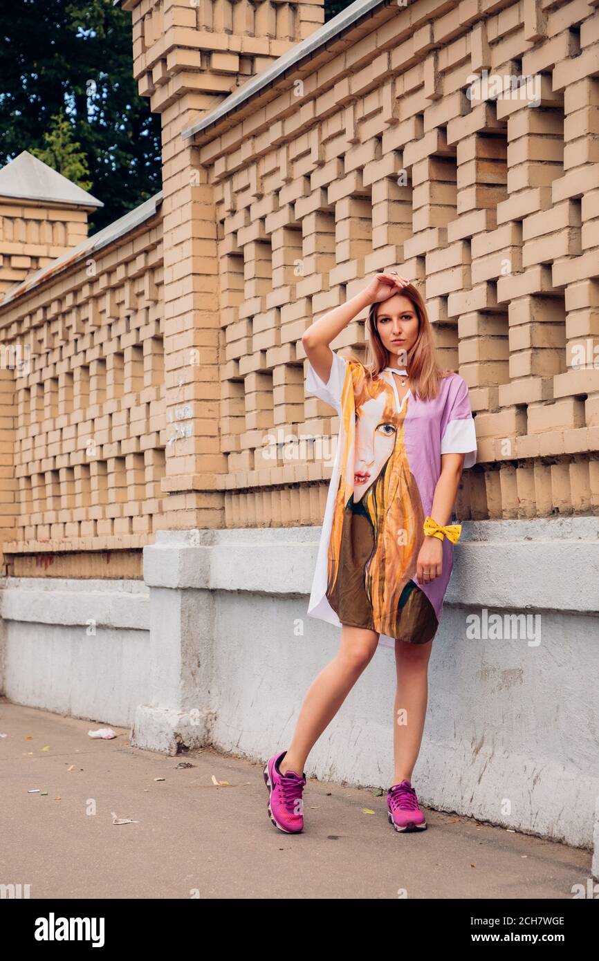 Pretty blonde girl is wearing bright pink sneakers and a colorful  dress with a face (similar to her face) on it; she stands in front of a stone wall Stock Photo