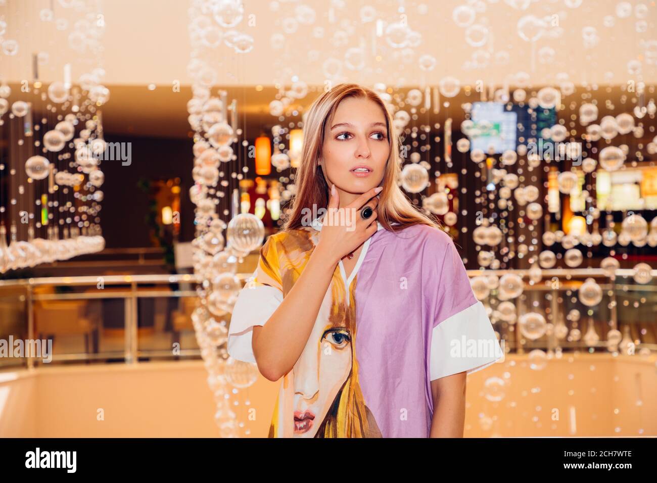 Girl in a colorful dress with a face (similar to hers) on it;she looks sidewards; in background glass balls are hanging from the ceiling as decoration Stock Photo