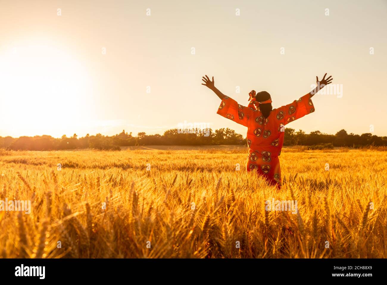 African woman in traditional clothes standing arms raised in field of barley or wheat crops at sunset or sunrise Stock Photo
