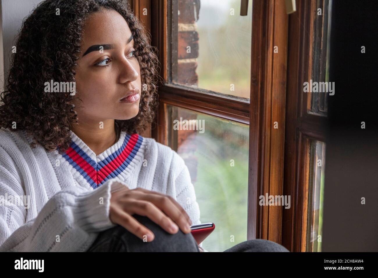 Beautiful mixed race African American girl teenager female young woman sad depressed or thoughtful looking out of a window using mobile cell phone Stock Photo