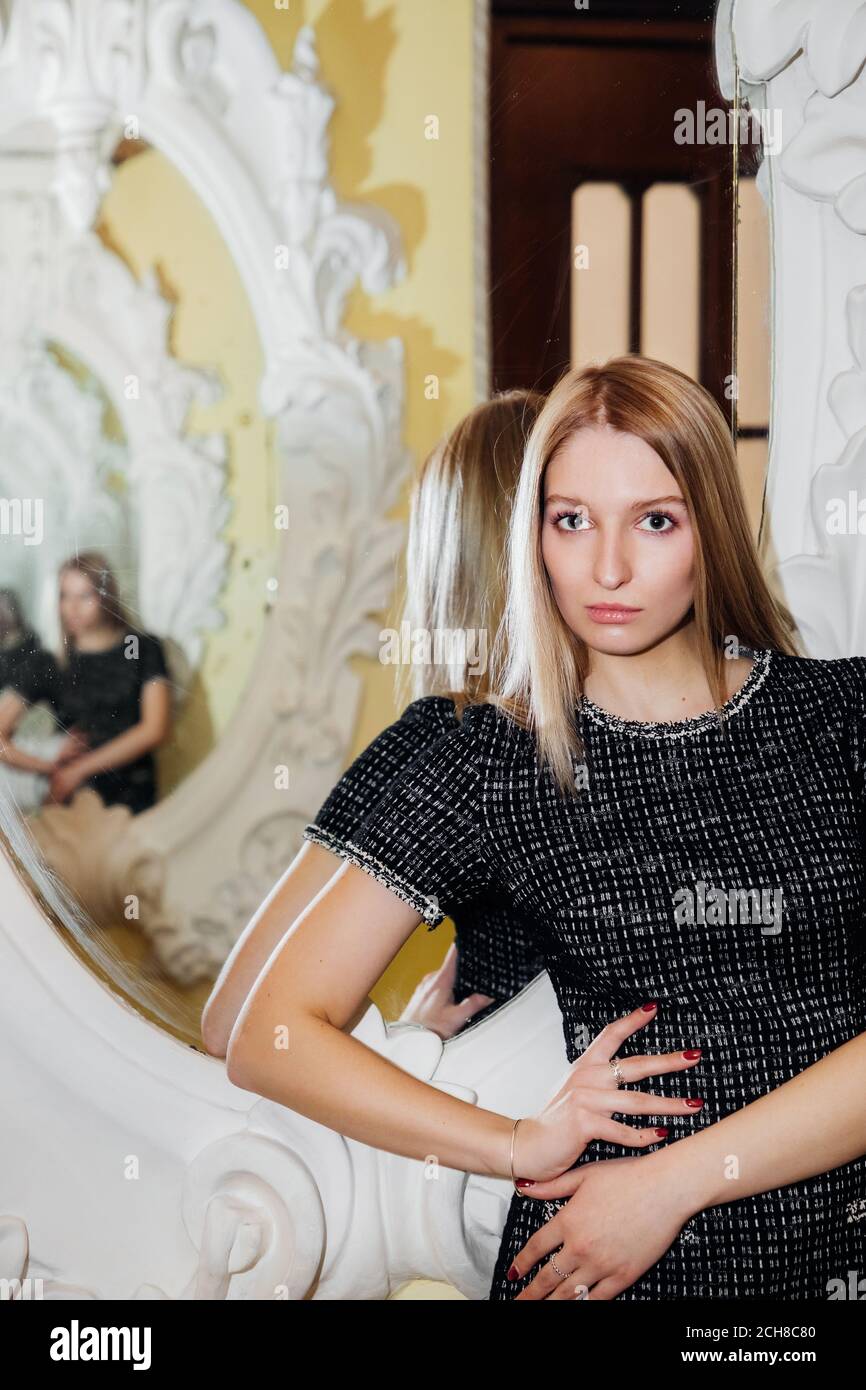 Pretty blonde girl is standing in front of a richly decorated mirror with multiple reflections; she is wearing a stylish dark dress with short sleeves Stock Photo