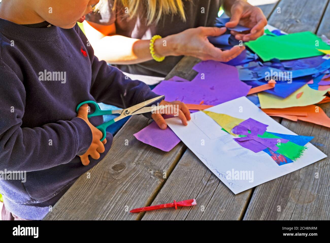Young child aged 3 learning to cut paper with scissors sitting outside on a table with colourful art paper in Carmarthenshire Wales UK  KATHY DEWITT Stock Photo