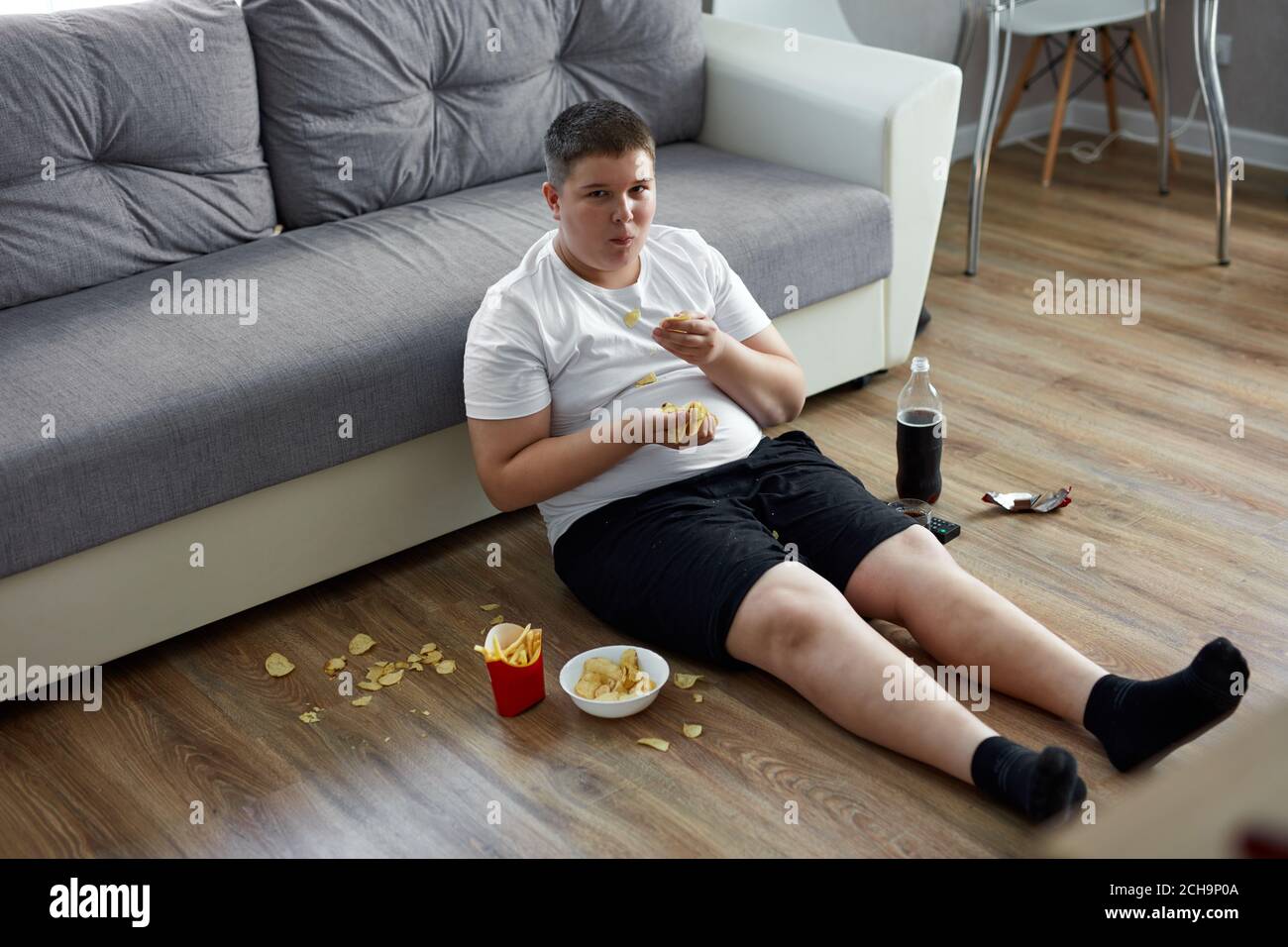 overweight fat boy eat junk food while watching tv alone at home, sit on the floor with french fries, carbonated drink, chips. enjoy unhealthy lifesty Stock Photo