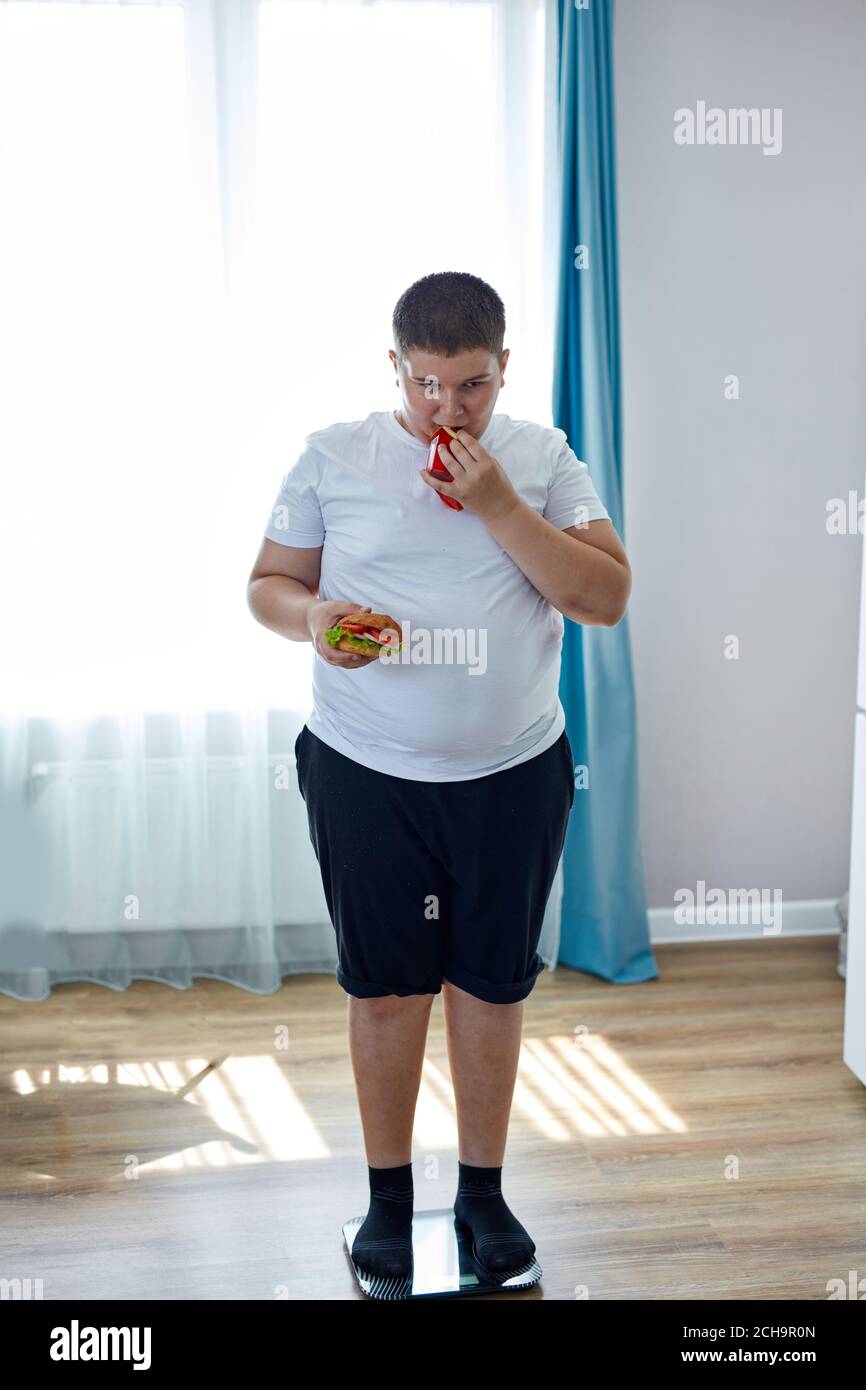 an overweight child suffer from having bad nutrition , stand on scales, look at weight and hold junk food in hands. passive unhealthy lifestyle concep Stock Photo