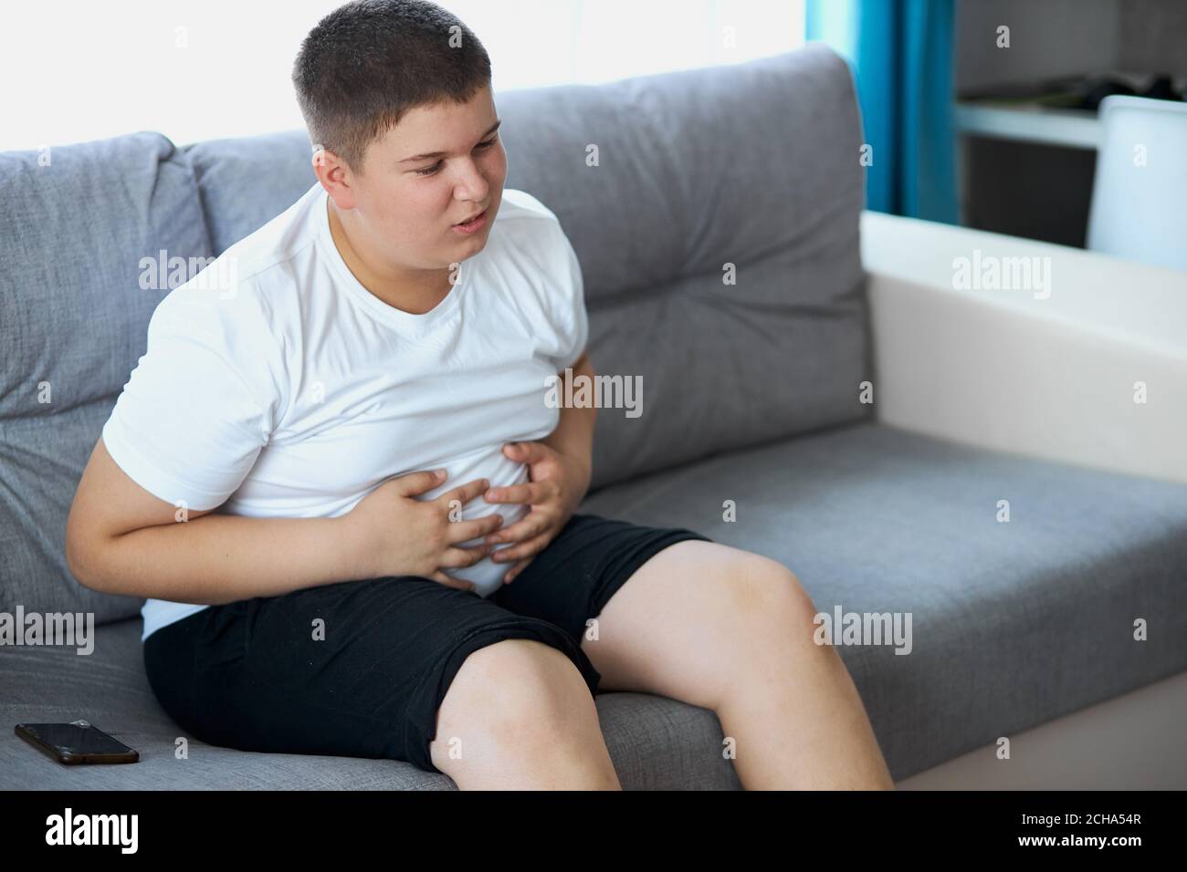 fat teenager boy has stomach ache after junk food, sit on sofa holding