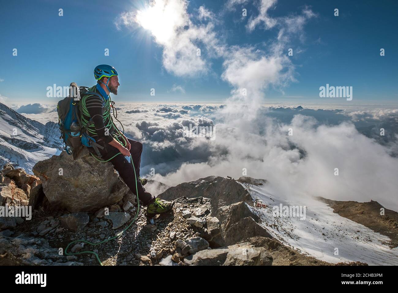 Bearded Climber in a safety harness, helmet, and on body wrapped climbing rope with sitting at 3600m altitude on a cliff and looking at  picturesque c Stock Photo