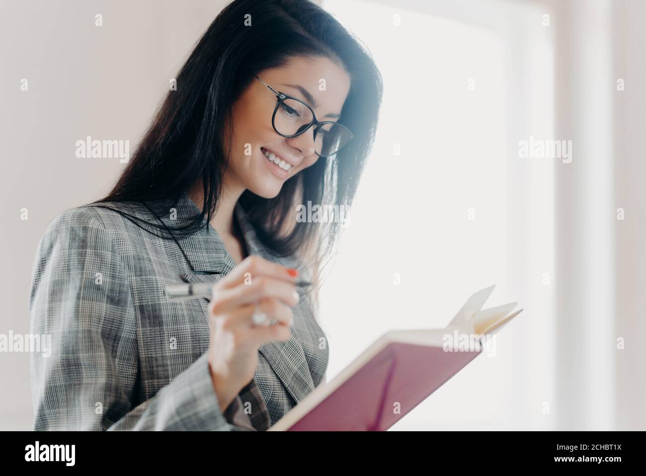 Sideways shot of pleasant looking woman has happy expression makes notes in planner