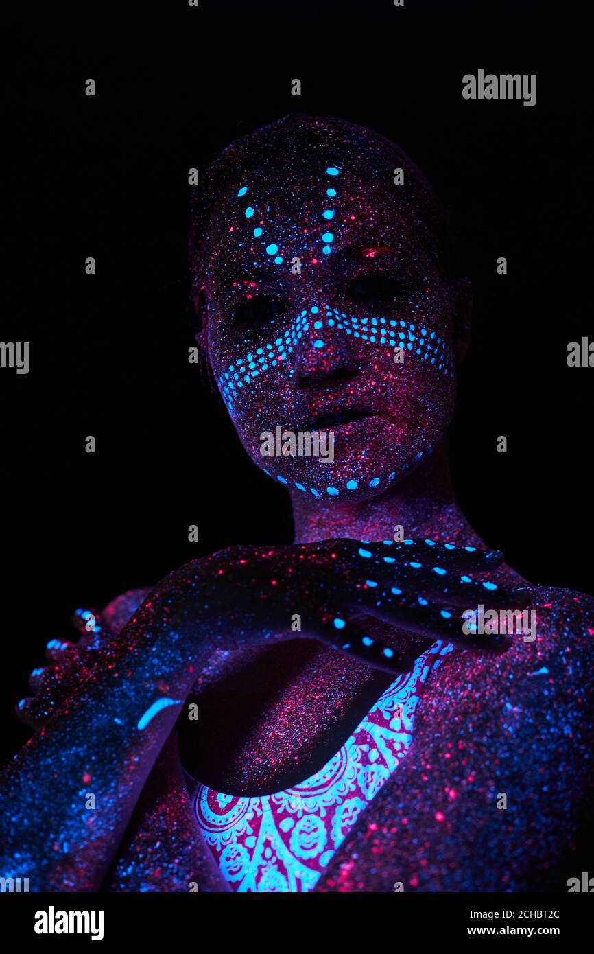 young black woman with fluorescent prints on skin, cosmic paint glowing on  neon lights, black background in studio. female with body art closing half  of face, touching. beauty, fashion concept Stock Photo
