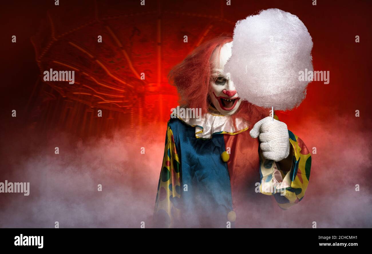 an eerie clown in front of the abstract background of a fun fair tries to lure with cotton candy Stock Photo