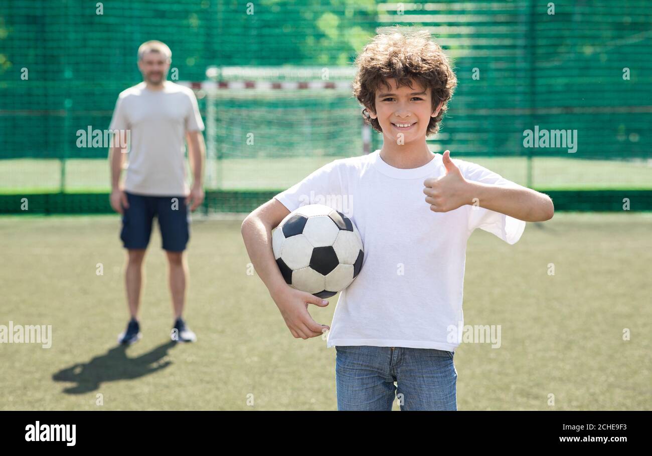 Boy holding soccer ball on the field, showing thumbs up Stock Photo