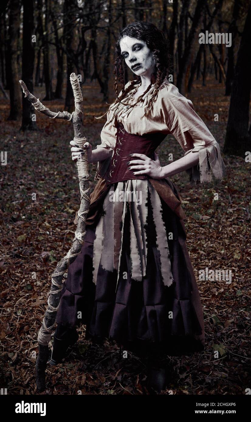Halloween theme: ugly scary voodoo witch with stick. Portrait of the evil sorceress in dark forest. Zombie woman (undead) Stock Photo