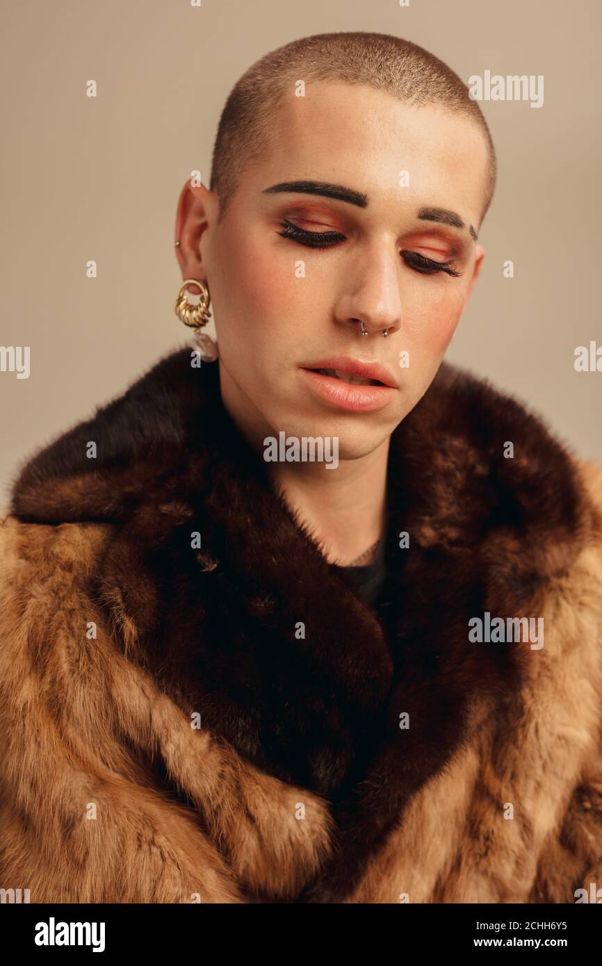 Close-up of androgynous male wearing makeup, earring and fur coat. Fashionable gay man against beige background. Stock Photo