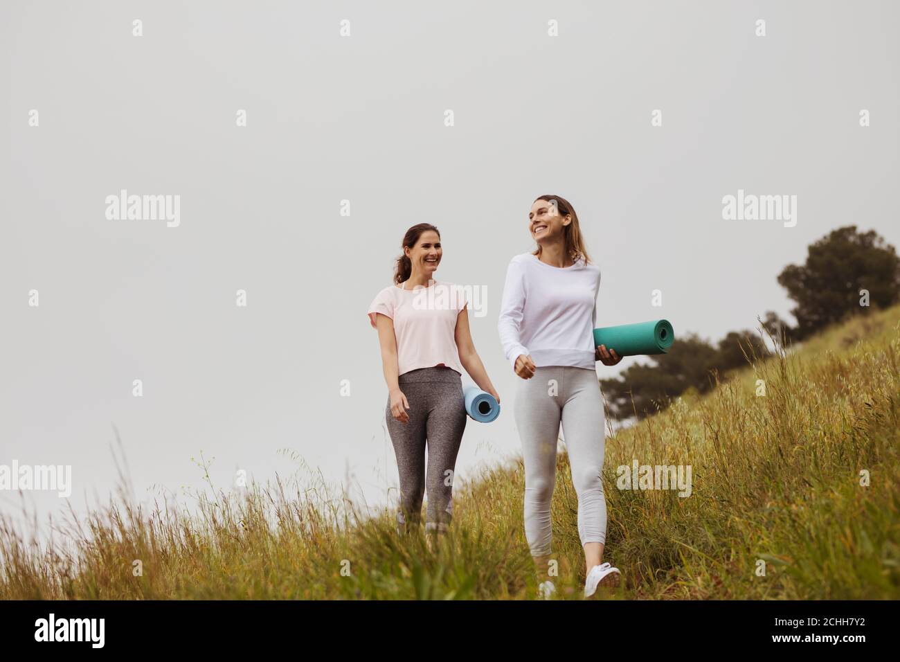 Two cheerful women friends walking with yoga mats in the countryside. Smiling women going for workout in the morning. Stock Photo