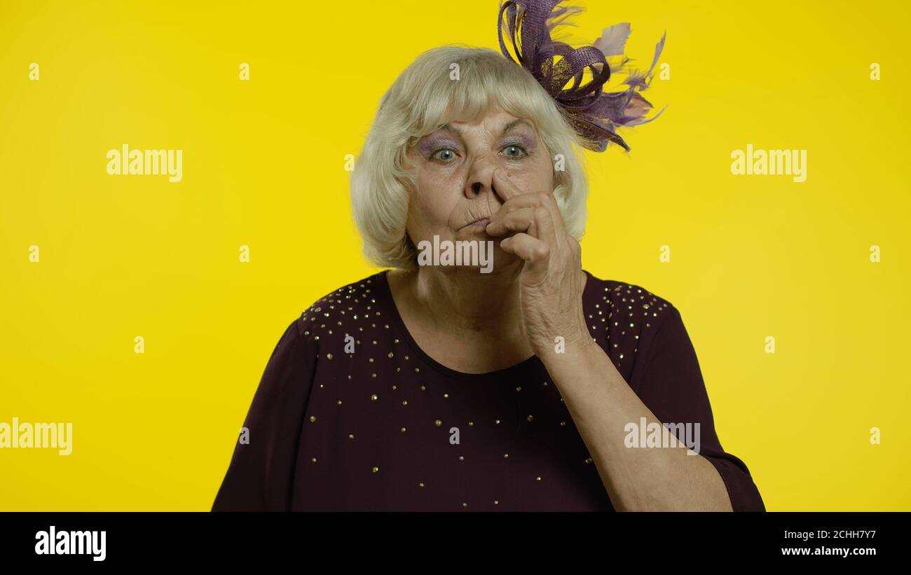 Funny stupid senior old woman picking nose with silly brainless humorous expression, removing boogers, uncultured habit, bad manners. Elderly stylish lady grandma on yellow background Stock Photo