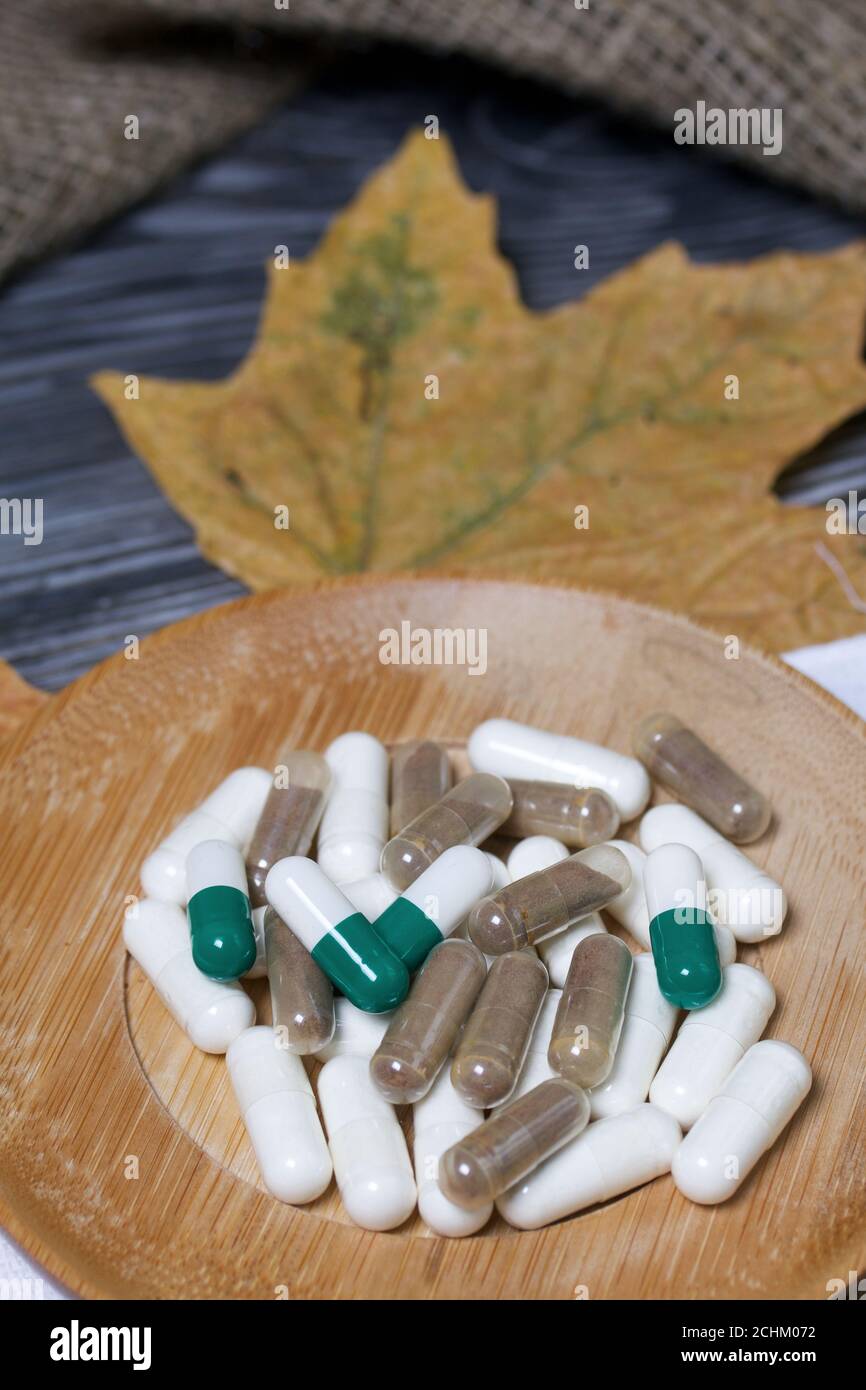 A handful of pills of different colors on a wooden saucer. Among the dried up maple leaves. On a pine board surface. Stock Photo