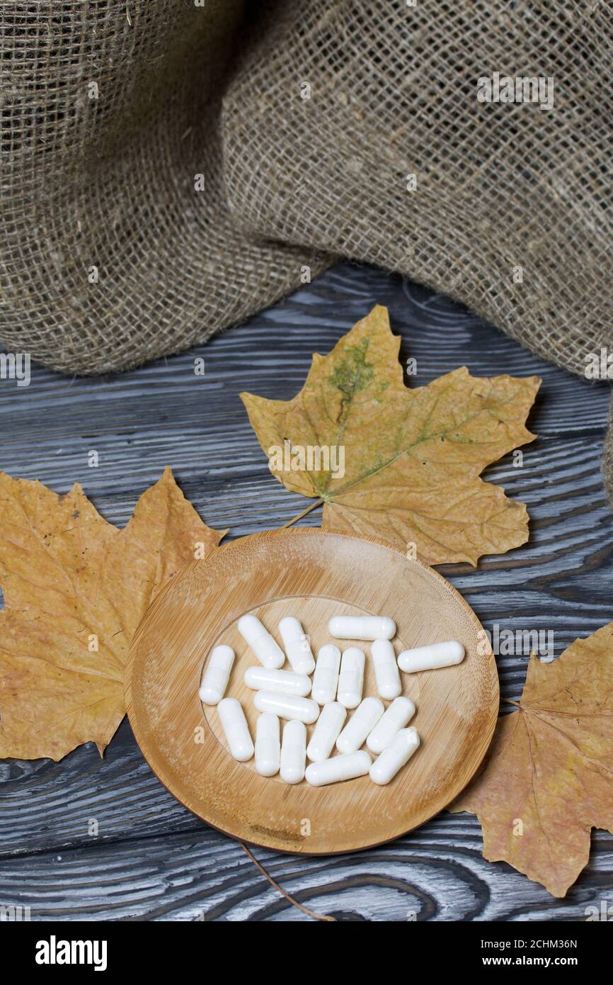 A handful of white pills on a wooden saucer. Among the dried up maple leaves. On a pine board surface. Stock Photo
