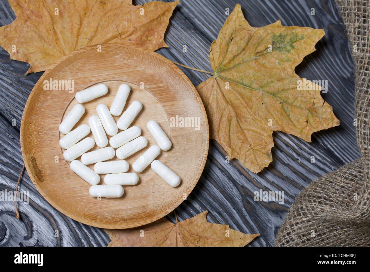 A handful of white pills on a wooden saucer. Among the dried up maple leaves. On a pine board surface. Stock Photo