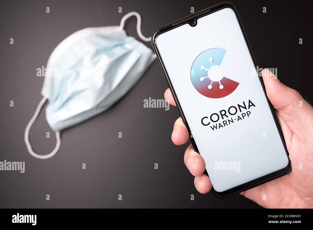 2020-09-14 Hamburg, Germany: hand holding smartphone with German COVID-19 contact tracing app against dark background with disposable face mask Stock Photo