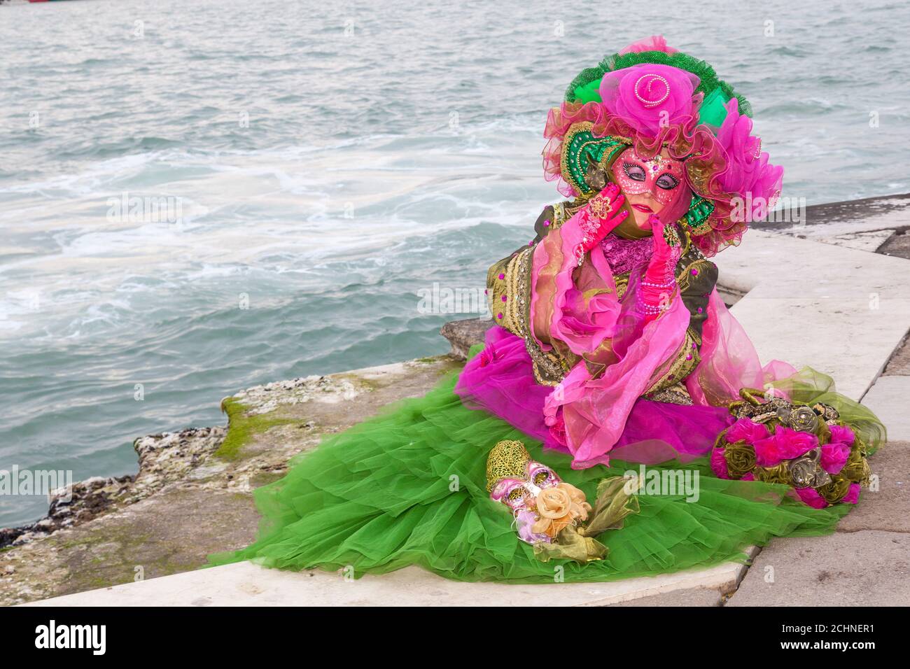 Venice, Veneto, Italy - woman in colorful green and pink costume posing at the edge of the water at the  lagoon at the Venice Carnival Stock Photo