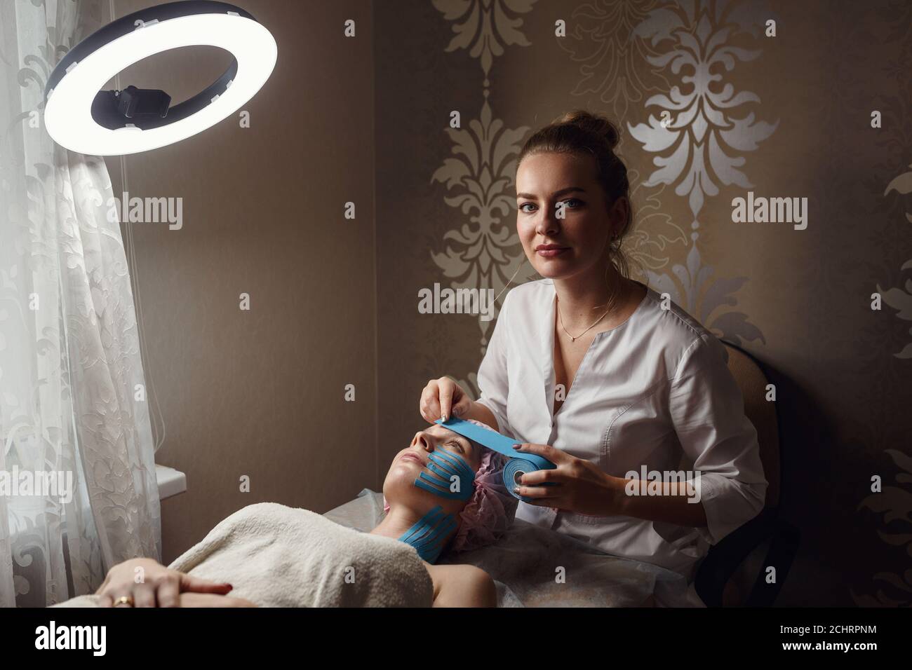 female cosmetician doing the taping of the face of the patient lying on the desk in the home. Physiotherapy and cosmetology procedure. Method of non-s Stock Photo