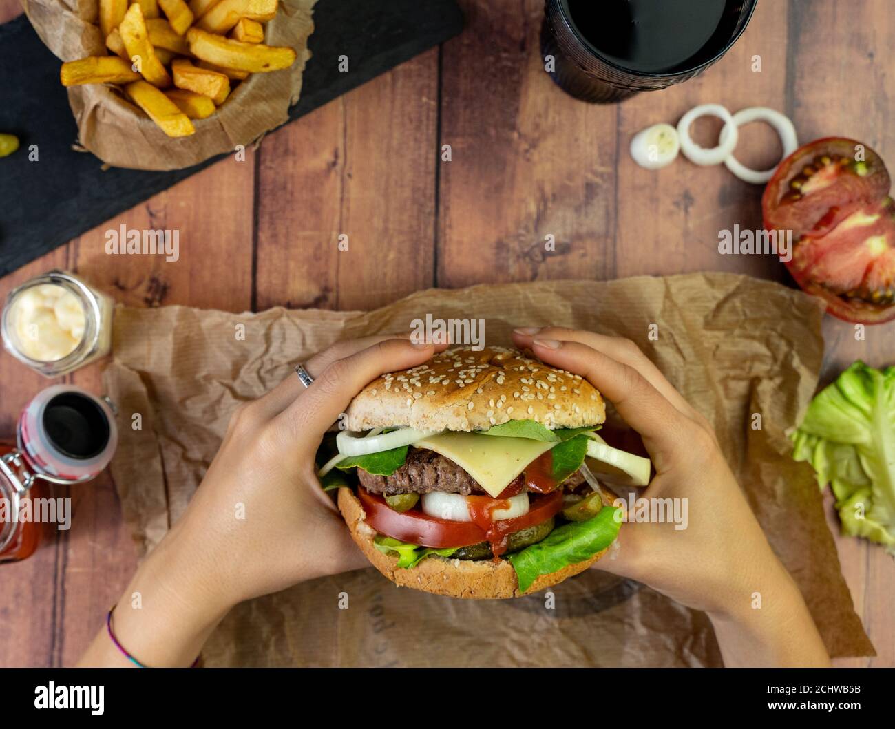 Person holding a burger with fries, onion rings and tomato on wooden table Stock Photo