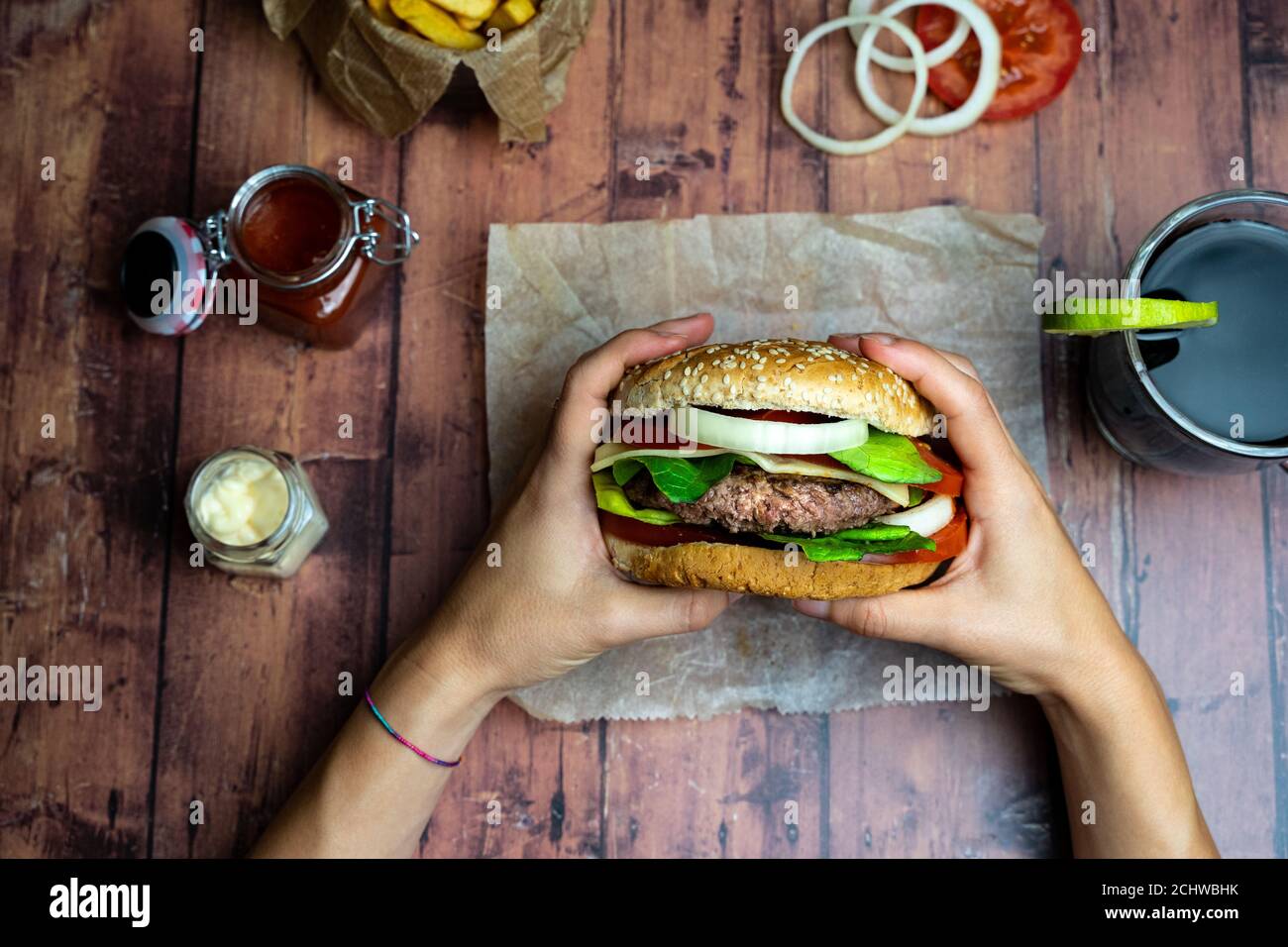 Person holding a burger with fries, onion rings and tomato on wooden table Stock Photo
