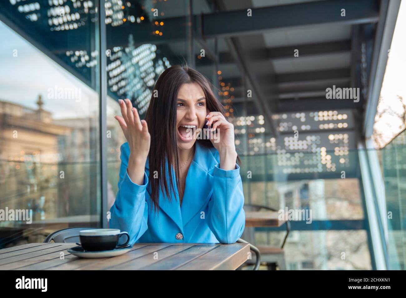 Portrait angry young woman in rage screaming while talking on mobile phone hand in air in frustration sitting at a table on balcony at home apartment Stock Photo