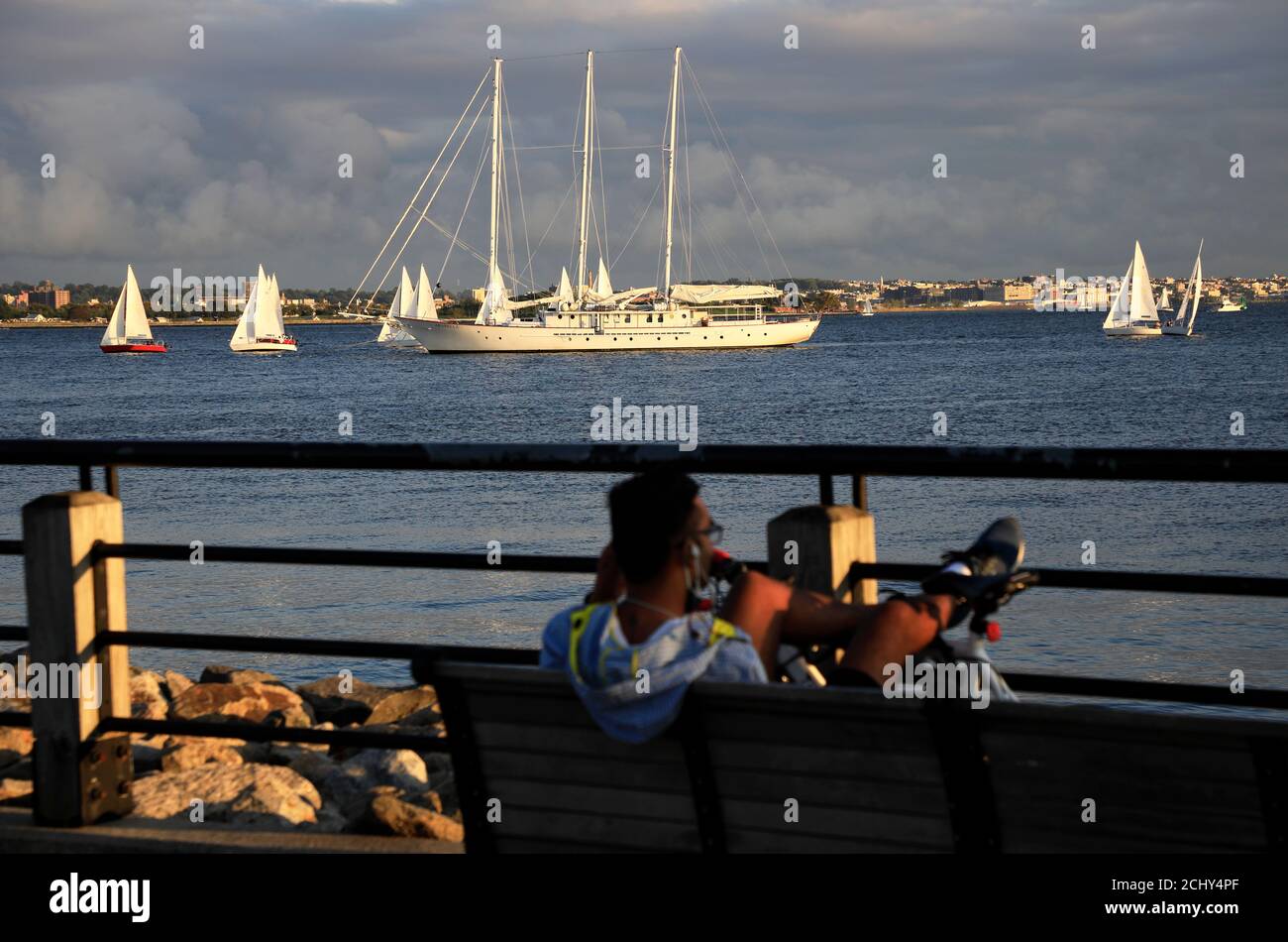 Man relaxing on a bench at Hudson River Waterfront Walkway with sailboats in the water of Upper New York Bay in background.Liberty State Park NJ.USA Stock Photo
