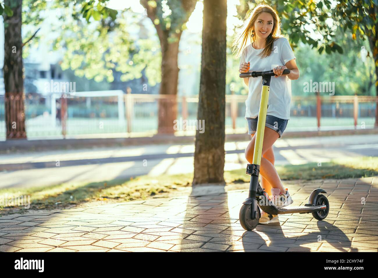 Smiling woman rides electric scooter or e-scooter in city park at sunset. Female using electric transport in urban park Stock Photo