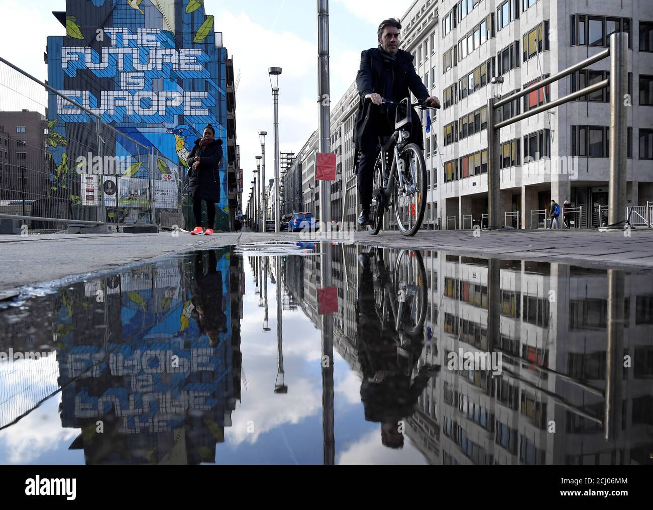 People are reflected in a puddle as they pass by a mural near the EU headquarters in Brussels, Belgium March 20, 2019. REUTERS/Toby Melville Stock Photo