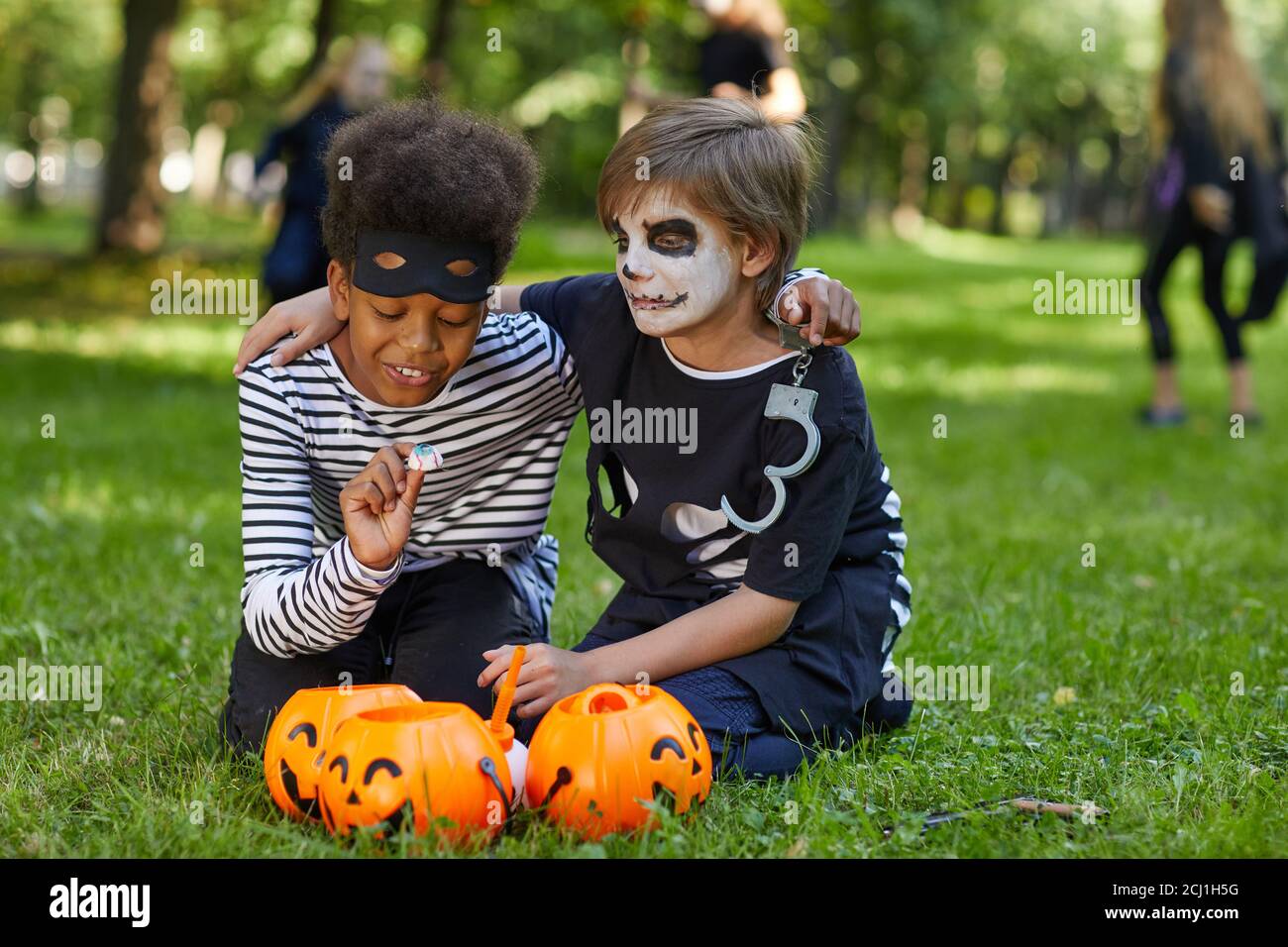 Full length portrait of two boys wearing Halloween costumes sitting on green grass outdoors with candy buckets, copy space Stock Photo