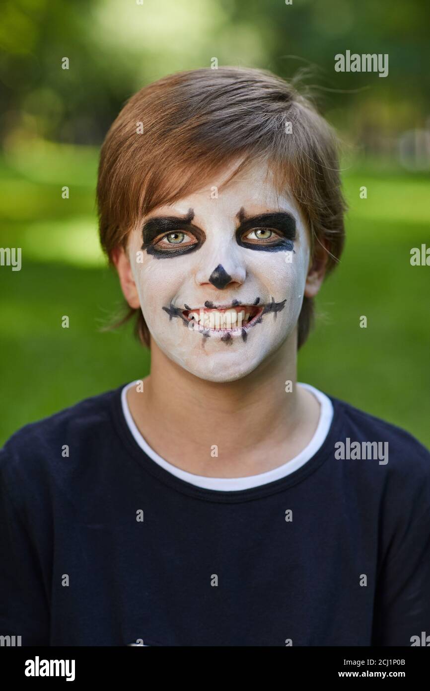 Head and shoulders portrait of smiling teenage boy wearing Halloween costume with face paint and looking at camera while posing outdoors Stock Photo