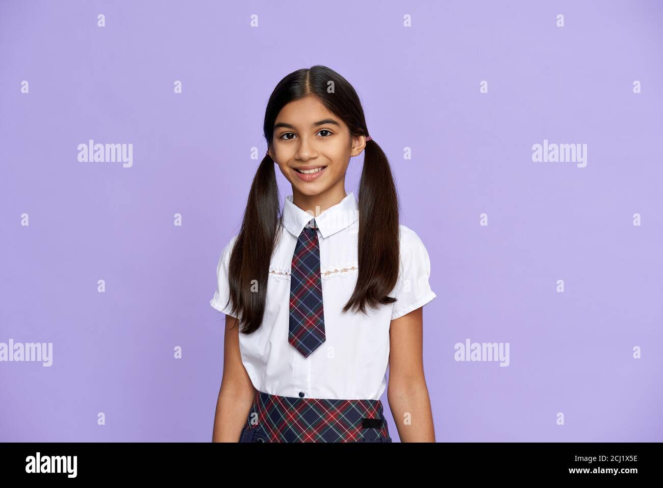Smiling indian kid girl wear school uniform stand on lilac background, portrait. Stock Photo