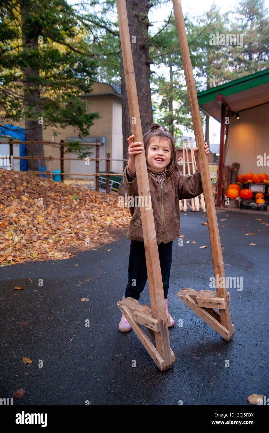 Small girl trying stilts on a pavement. Halloween display in a background. Stock Photo