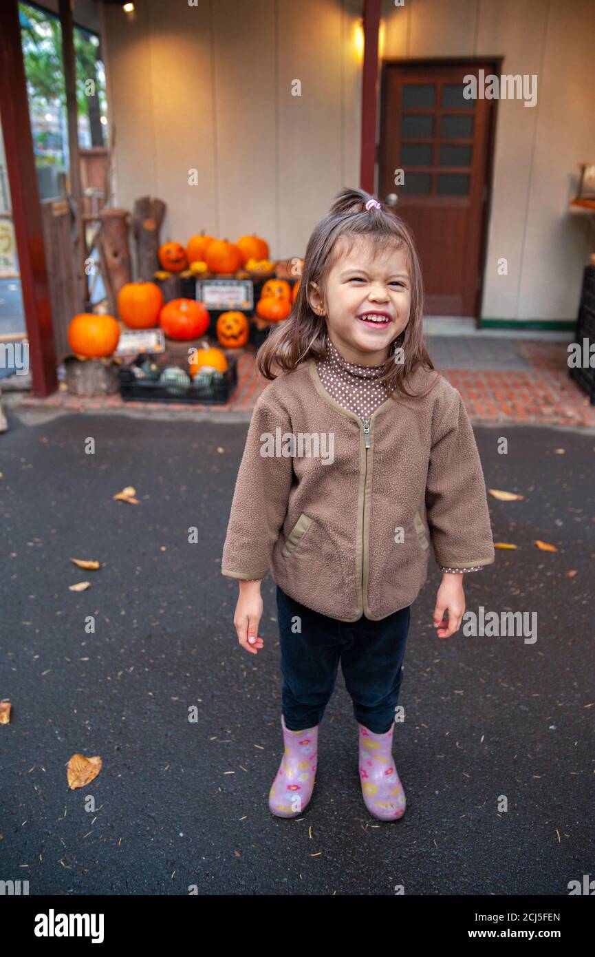Little girl in brown fleece jacket smiling and waiting for Halloween in front of an entrance with pumpkins. Stock Photo