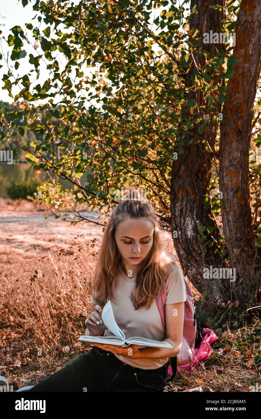 Young girl, student sitting outdoor with backpack and reading a book. Study from home. Cottagecore Stock Photo