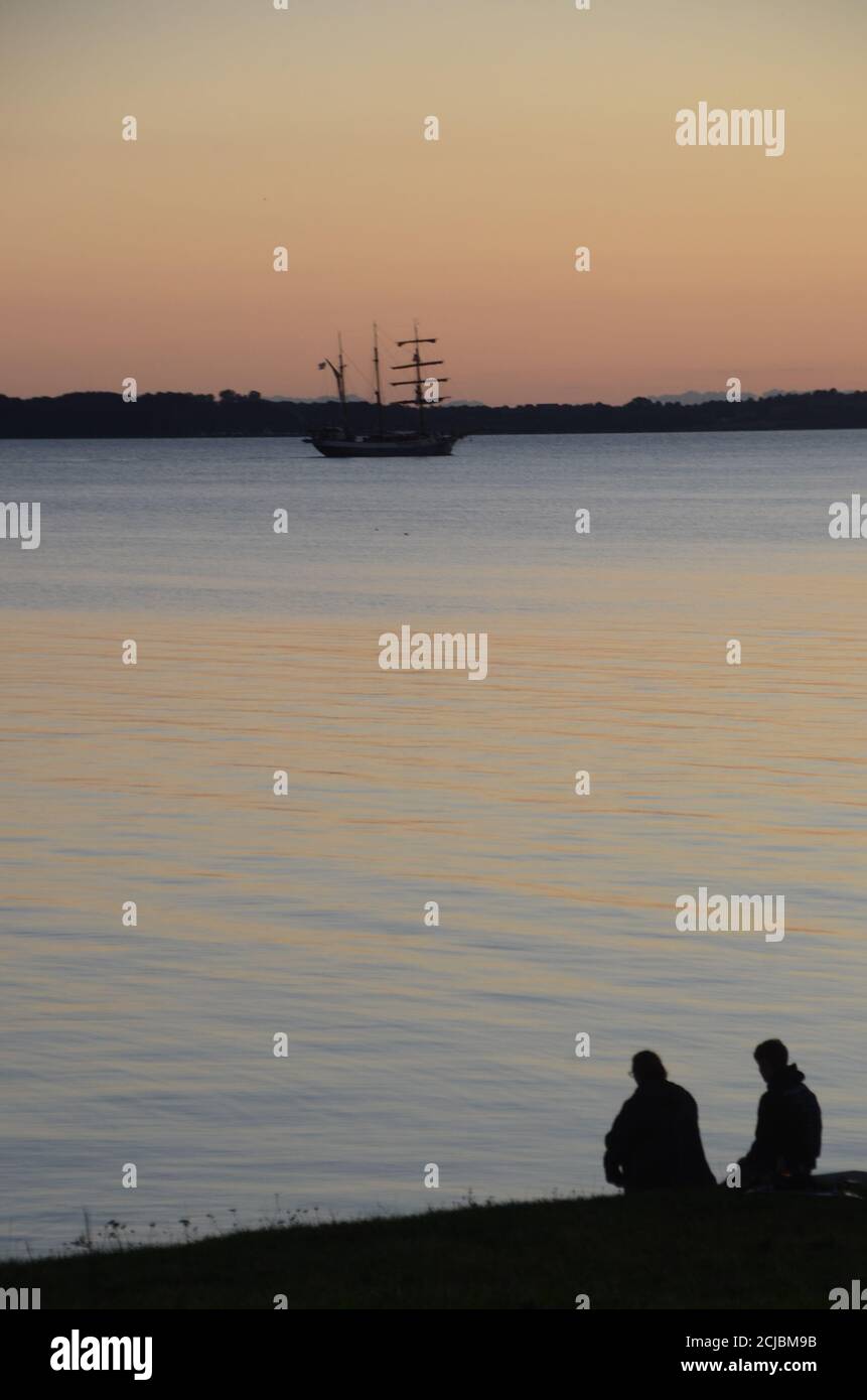 Old sail ship passes out on the sea while two persons in silhouette watch Stock Photo