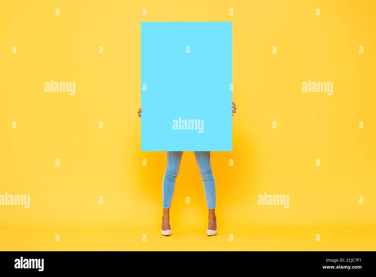 Woman holding and hiding behind empty blue placard in isolated studio yellow background Stock Photo