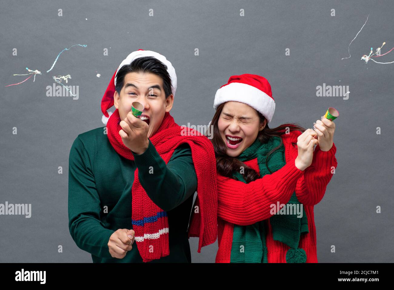 Cute excited Asian couple pulling party poppers celebrating Christmas in isolated studio gray background Stock Photo